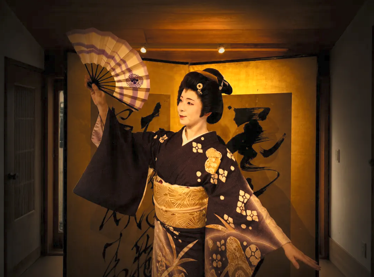 A woman dressed as a traditional Japanese geisha, holding a fan, standing in front of a gold and black artistic backdrop.