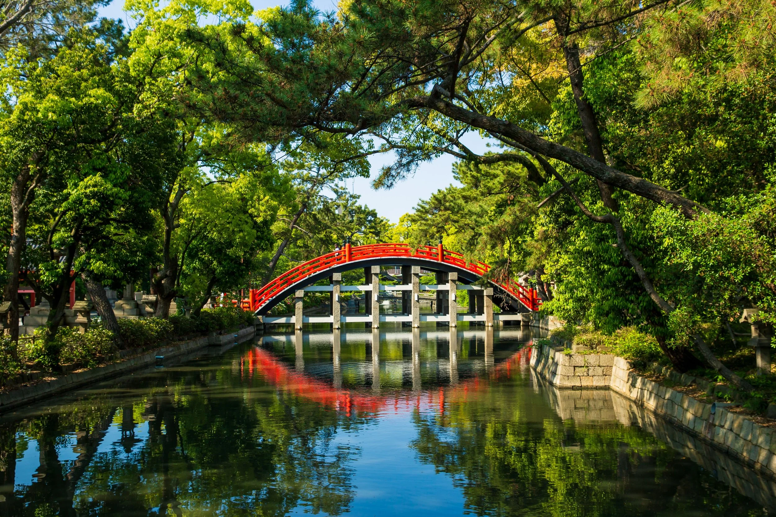 A scenic view of a red arched bridge crossing over a calm canal surrounded by lush green trees and foliage, reflecting in the water on a sunny day.
