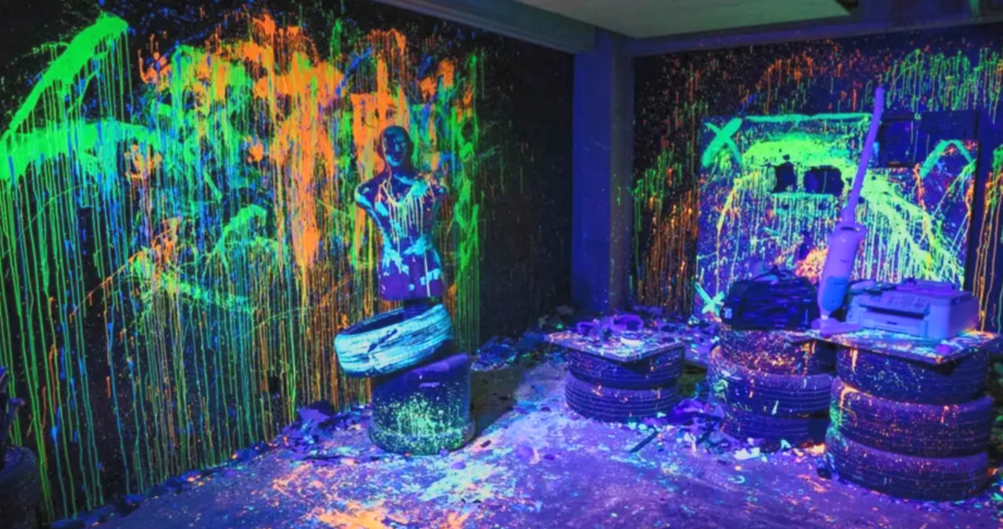 Room with black walls painted with neon paint, creating glowing graffiti art under blacklight, with artist supplies, and various tires stacked on the floor.