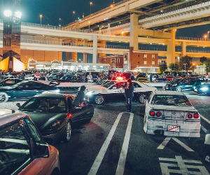 A crowded parking lot at night with numerous cars parked and some moving. There is a person walking among the cars and a police car with flashing lights. Overhead highways and streetlights are visible in the background.