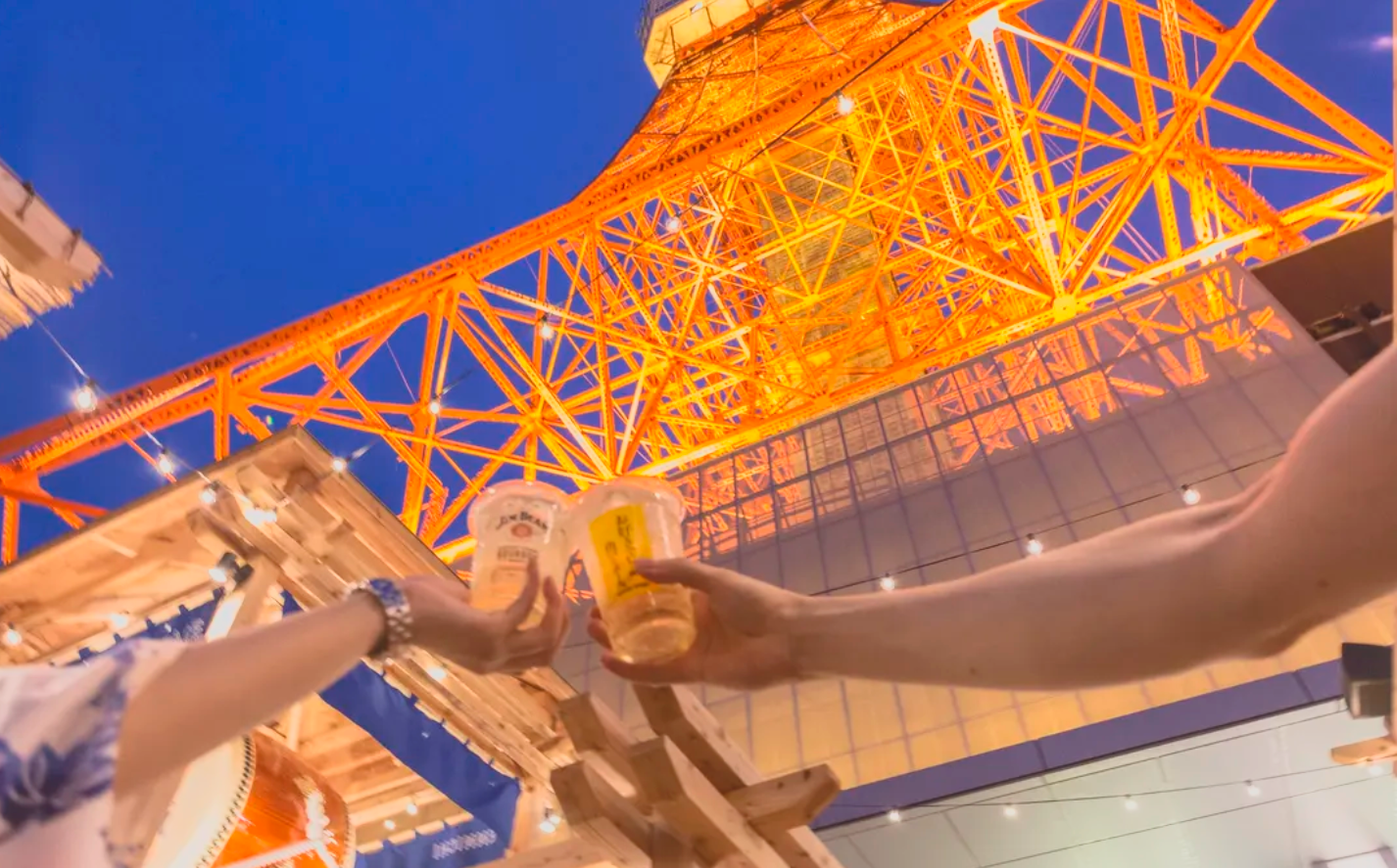 Two people holding drinks in front of Tokyo Tower illuminated in orange at night, viewed from below.