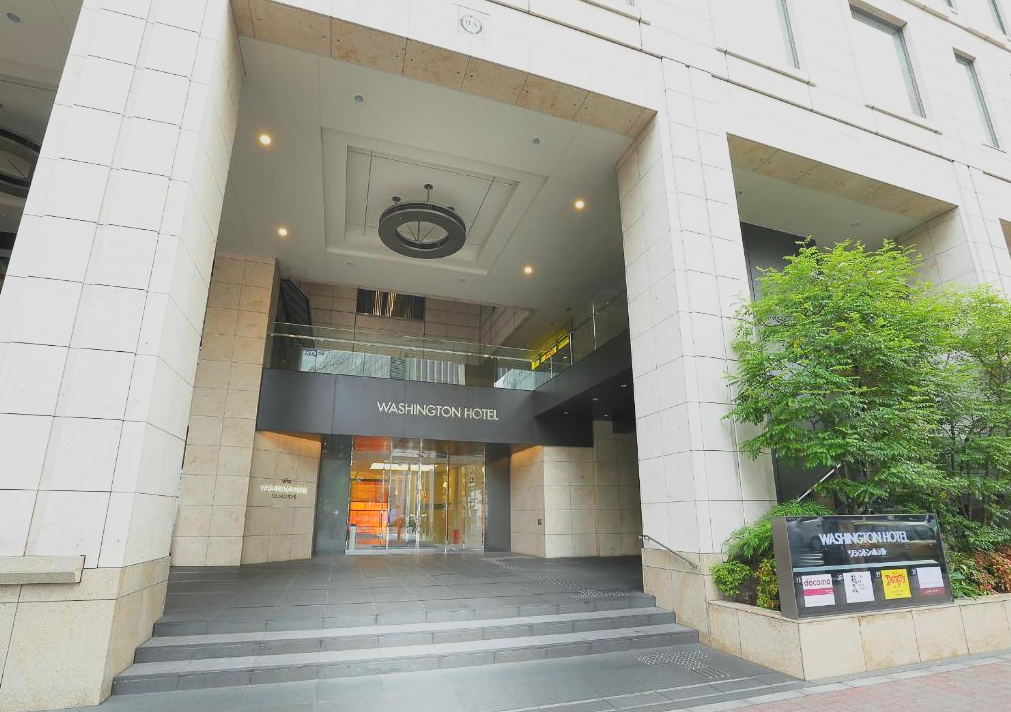 Entrance to the Washington Hotel with glass doors, black signage, and greenery to the right.