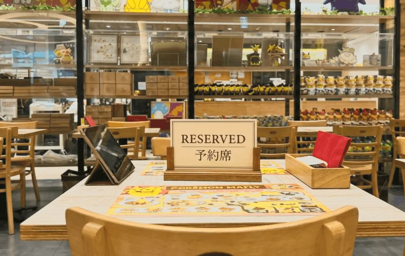 A table in a restaurant or cafe with a reserved sign, a menu, and a stylus; in the background, shelves filled with colorful desserts and decorative items.