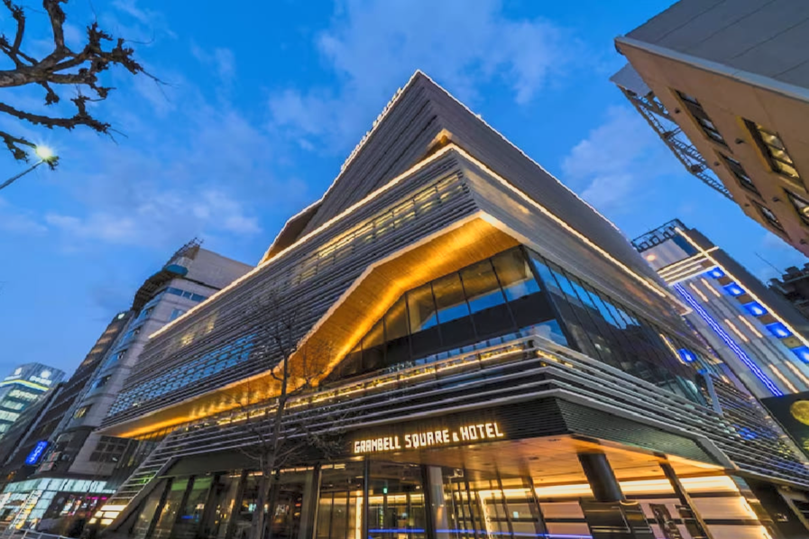 Modern hotel building with illuminated lines at dusk, labeled 'Gramell Square Hotel' at the entrance, surrounded by city buildings.