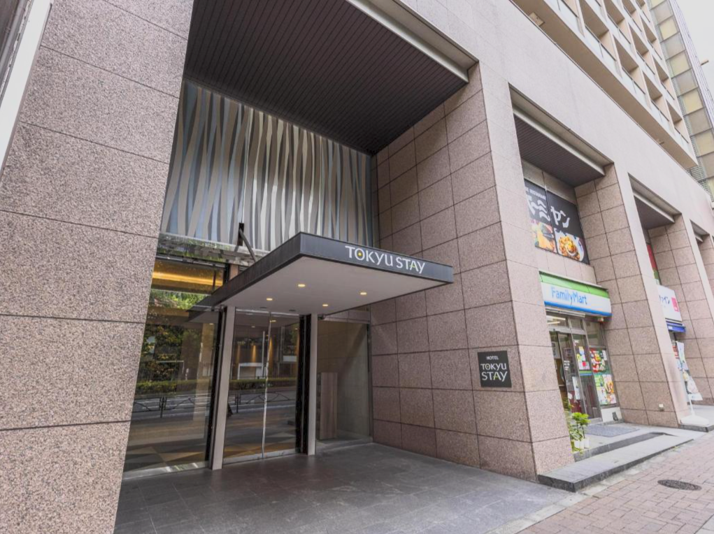 Close-up of the entrance to a hotel named 'Tokyu Stay,' with a sign displaying the hotel name, located on a city street with a FamilyMart convenience store adjacent to it.