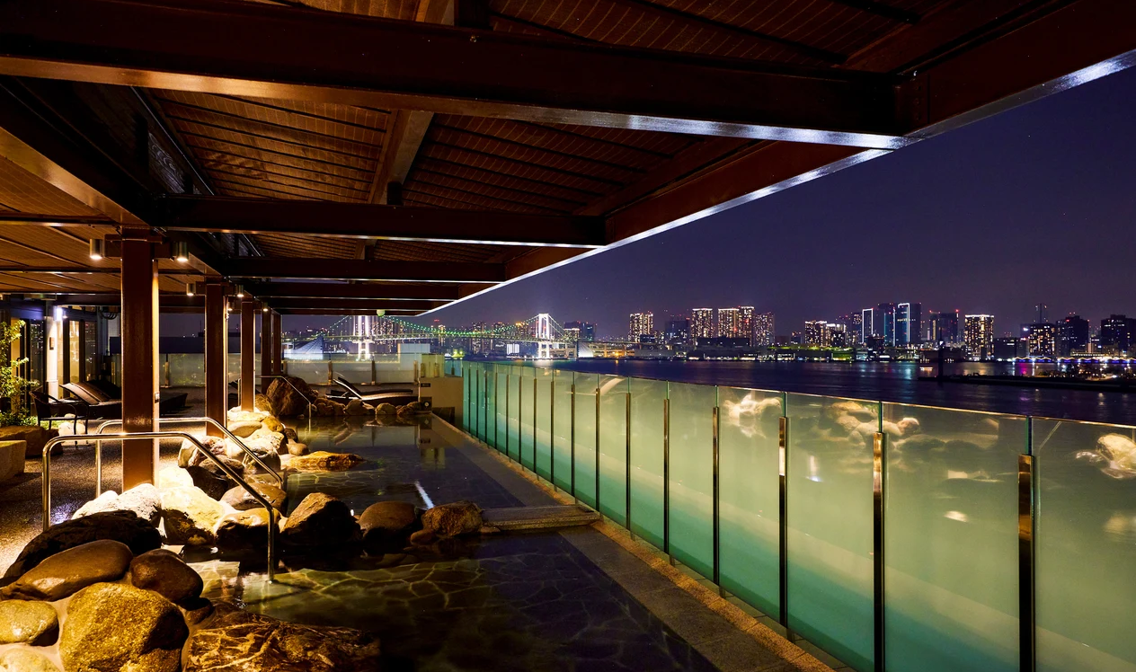 Nighttime view of a rooftop pool area overlooking a city skyline with illuminated buildings and a bridge in the distance.