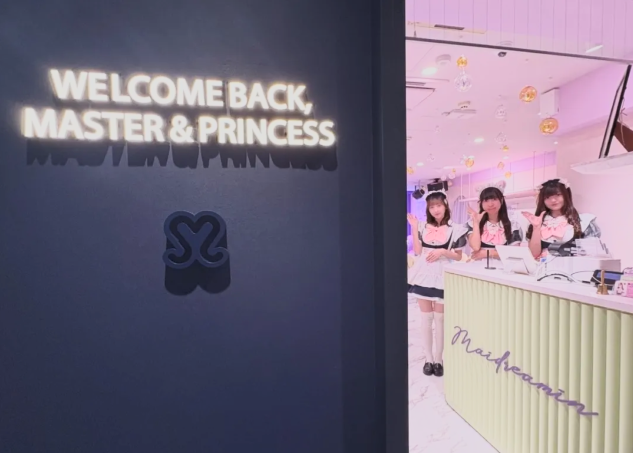A neon sign saying 'Welcome Back, Master & Princess' on a dark wall, with a small symbol beneath, next to a reception area where three young women dressed in maid costumes are standing behind a counter inside a pink-lit room decorated for Halloween.
