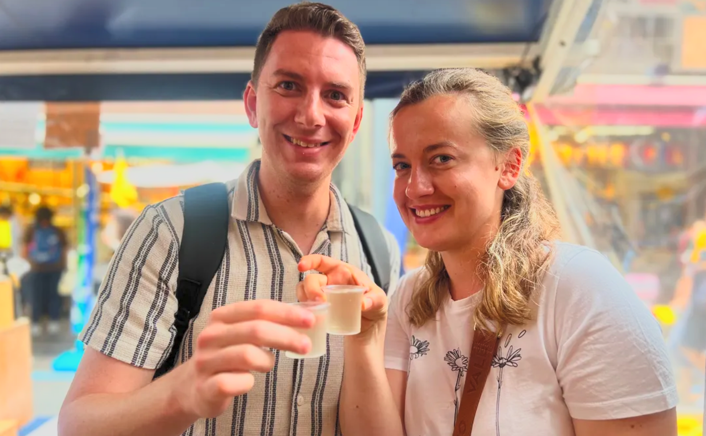 A smiling man and woman holding small cups in an amusement park or fair with colorful rides and attractions in the background.