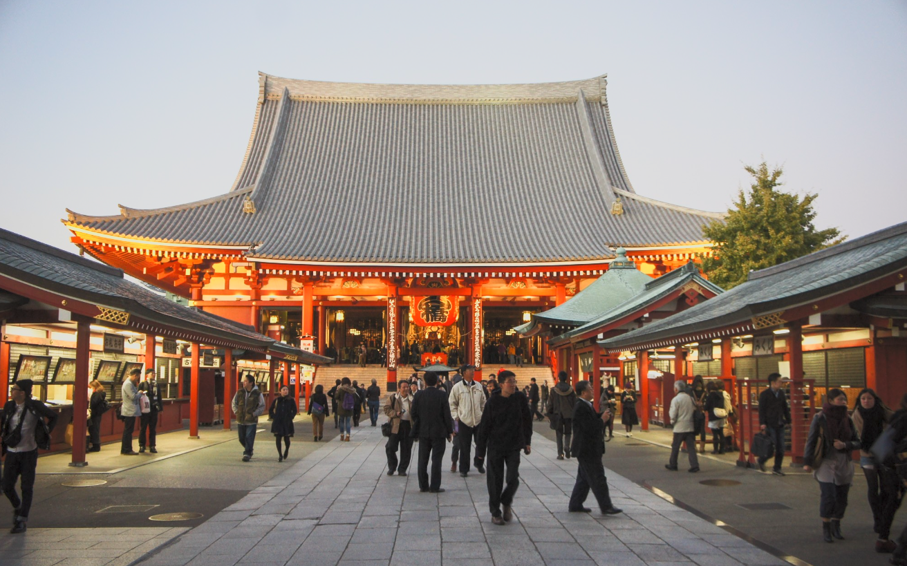 People walking toward and around a large traditional Japanese temple with orange and black coloring and a curved roof, at dusk.