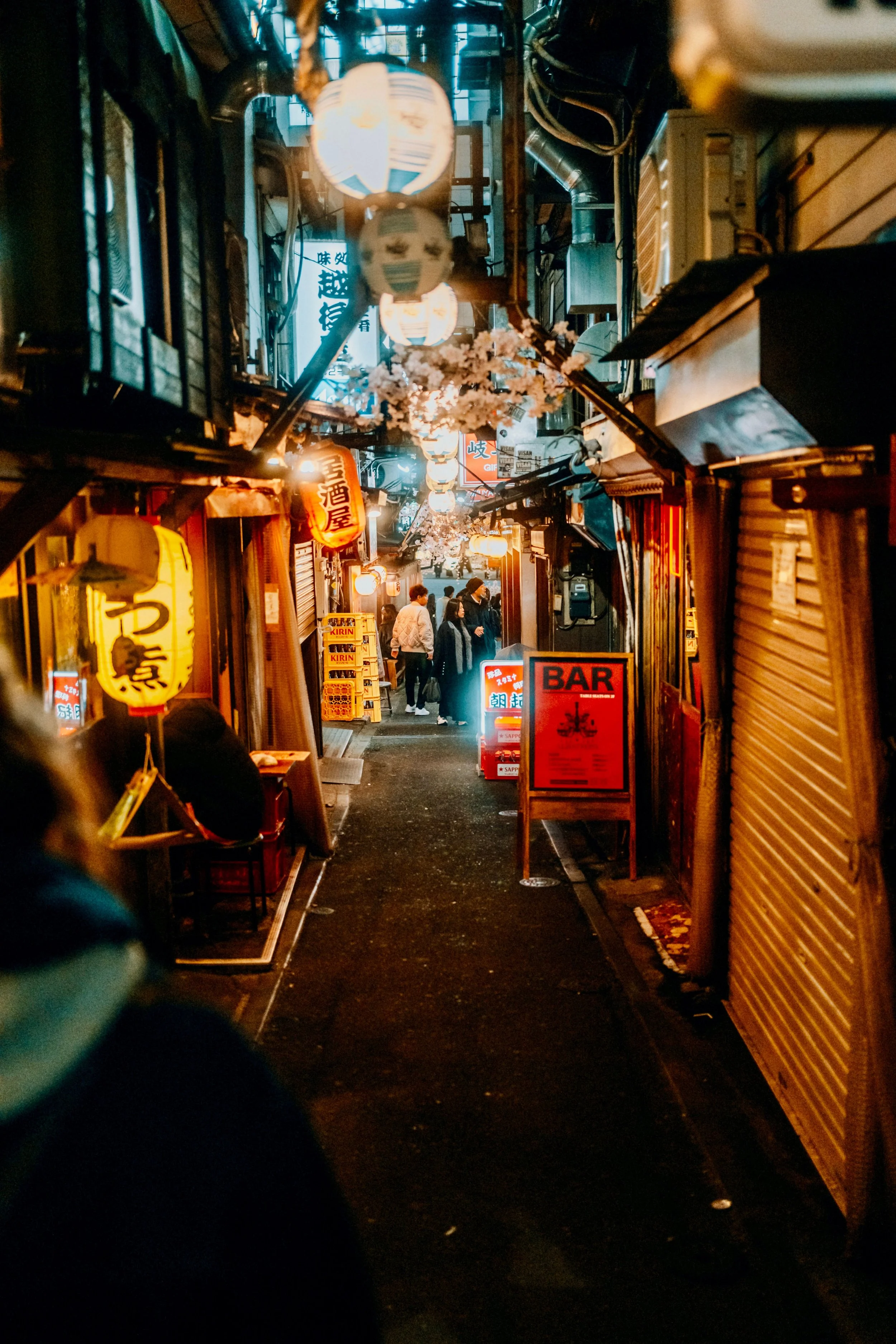 Nighttime street scene with narrow alley lined with closed shops with roll-up metal shutters. Hanging lanterns and string lights illuminate the street with warm colors. People are walking and sitting in small restaurants, some wearing jackets. Cherry blossom decorations are overhead, creating a festive atmosphere.