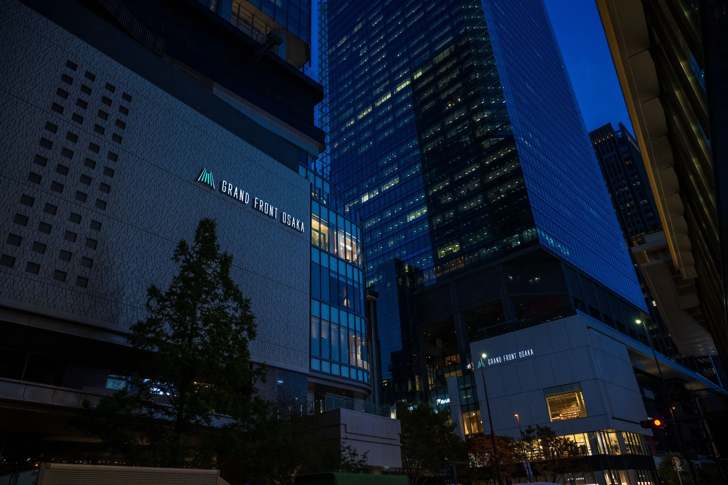 Nighttime view of modern glass skyscrapers in Osaka, Japan, with illuminated signs including "GRAND FRONT OSAKA" on a building, and city lights.