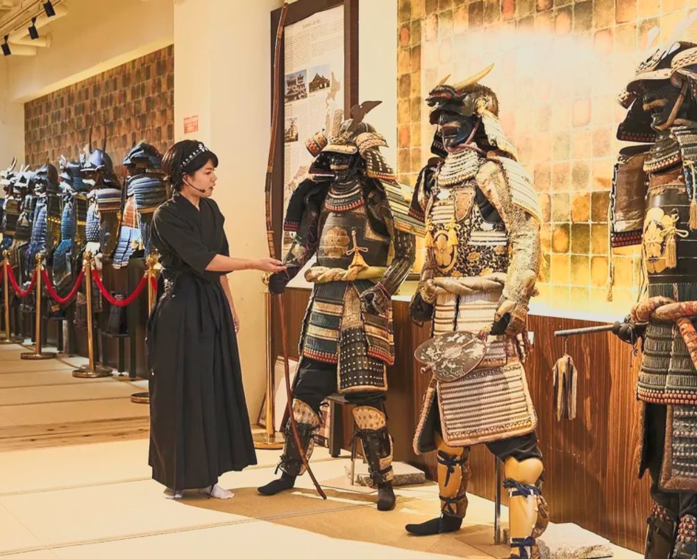 A woman in black kimono attire shows a sword to a display of traditional samurai armor in a museum.
