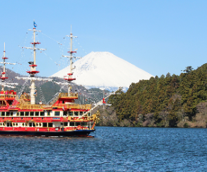 A red sailing ship near a body of water with snow-capped Mount Fuji in the background and a forested shoreline on the right.