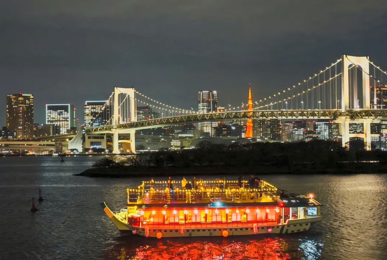 Night view of a lit-up boat on water with illuminated skyline and bridge in the background.
