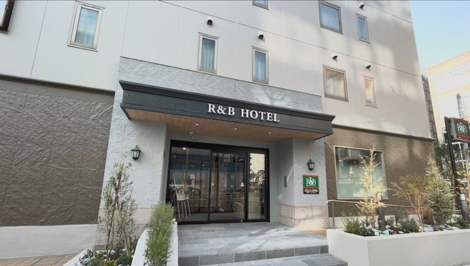 Entrance of the R&B Hotel with glass doors, a black sign above, outdoor plants on either side, and a welcome sign in the window.