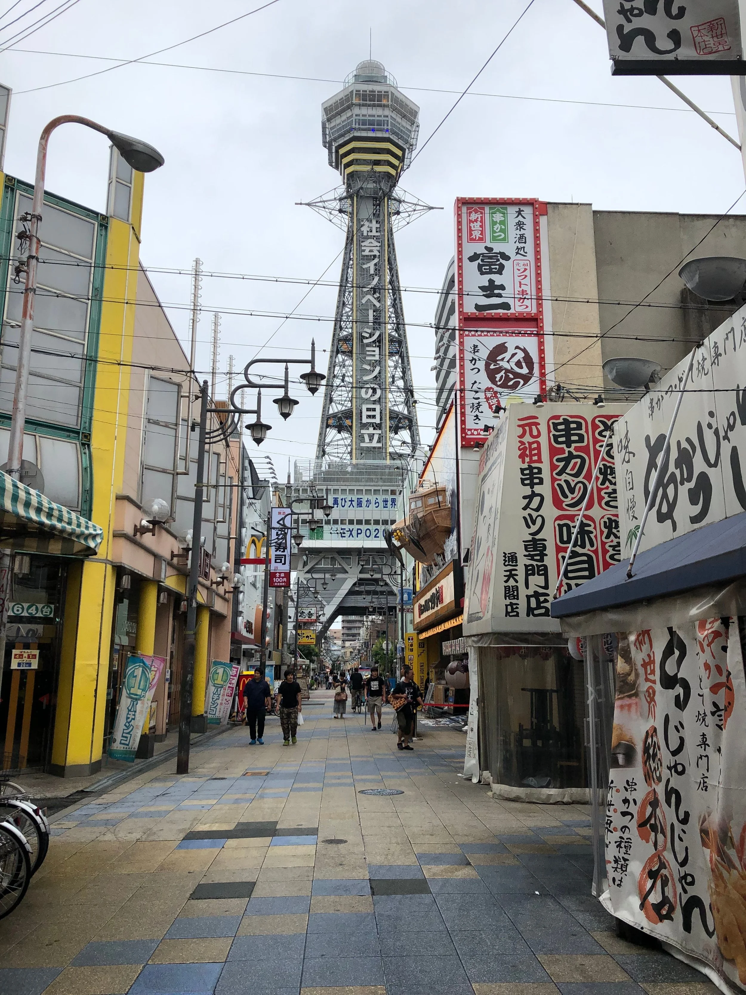 A busy street scene in Japan with a large tower in the background, shops and signs with Japanese writing, and pedestrians walking along the street.