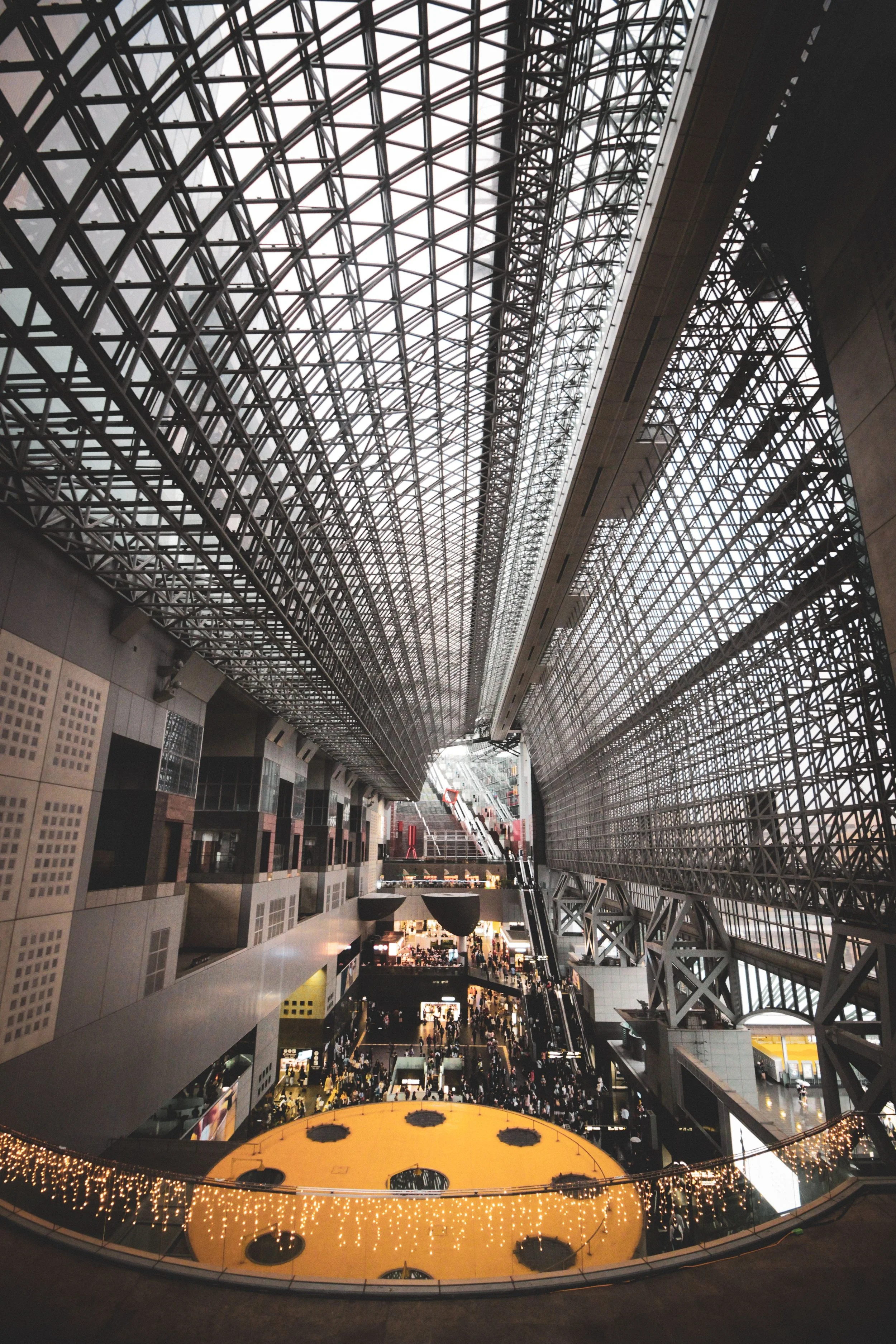 Interior of a large modern building with a high, arched glass ceiling and multiple levels of shops and walkways, filled with many people.