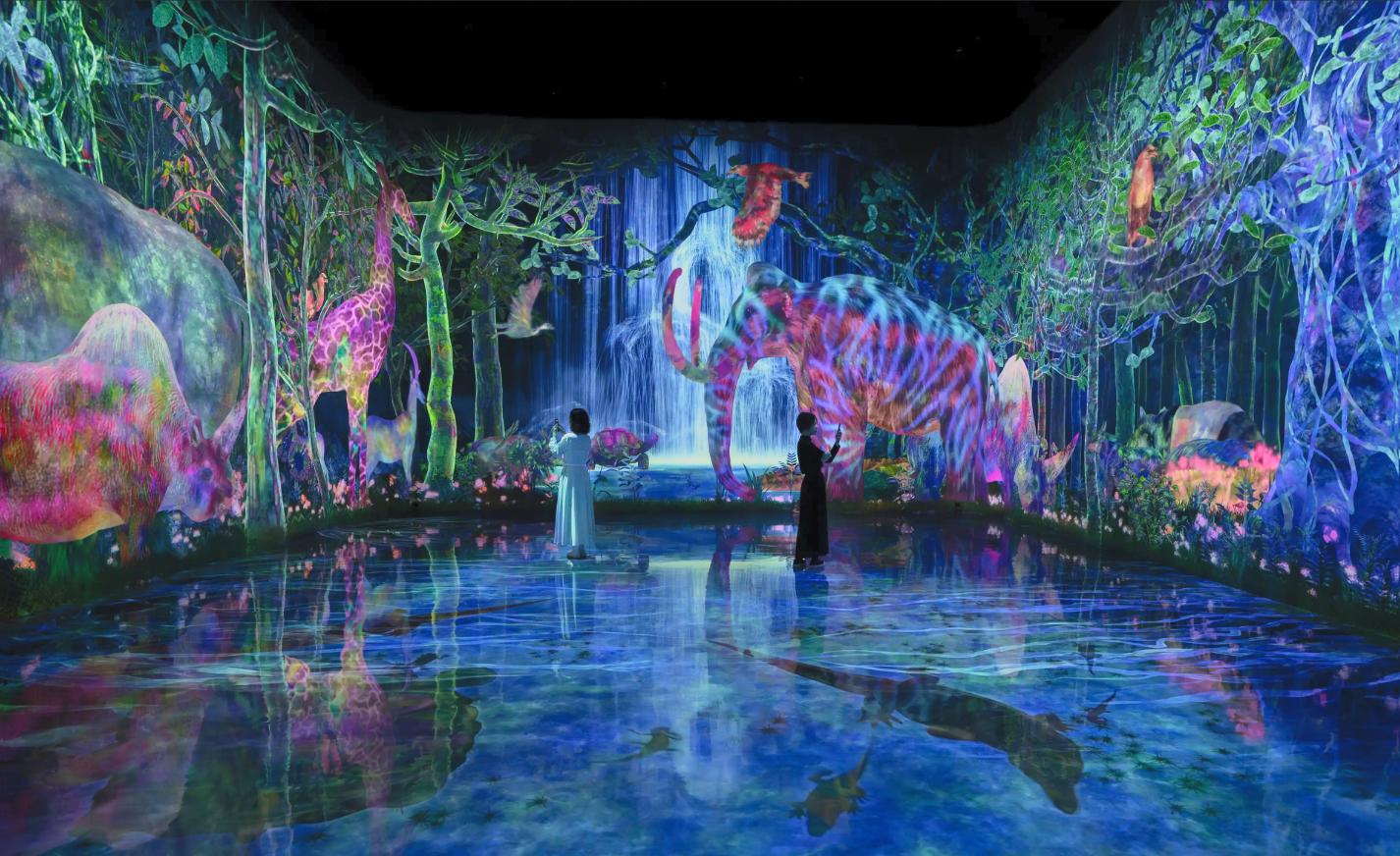 Interactive digital art installation depicting a colorful jungle scene with glowing animals, including an elephant, rhinoceros, giraffe, and birds, with cascading waterfalls in the background and two visitors observing the display.