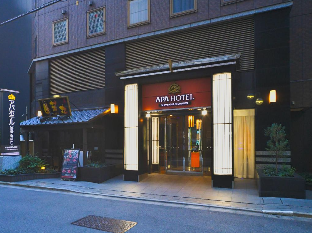 The entrance of APA Hotel, Shinbashi-Onarimon, with illuminated vertical signboards and a traditional Japanese roof structure on the left side.