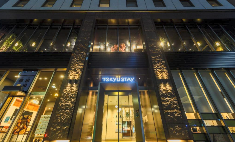 Tall modern building with illuminated windows and a sign that reads "Tokyu Stay" above the entrance, taken at dusk or night.