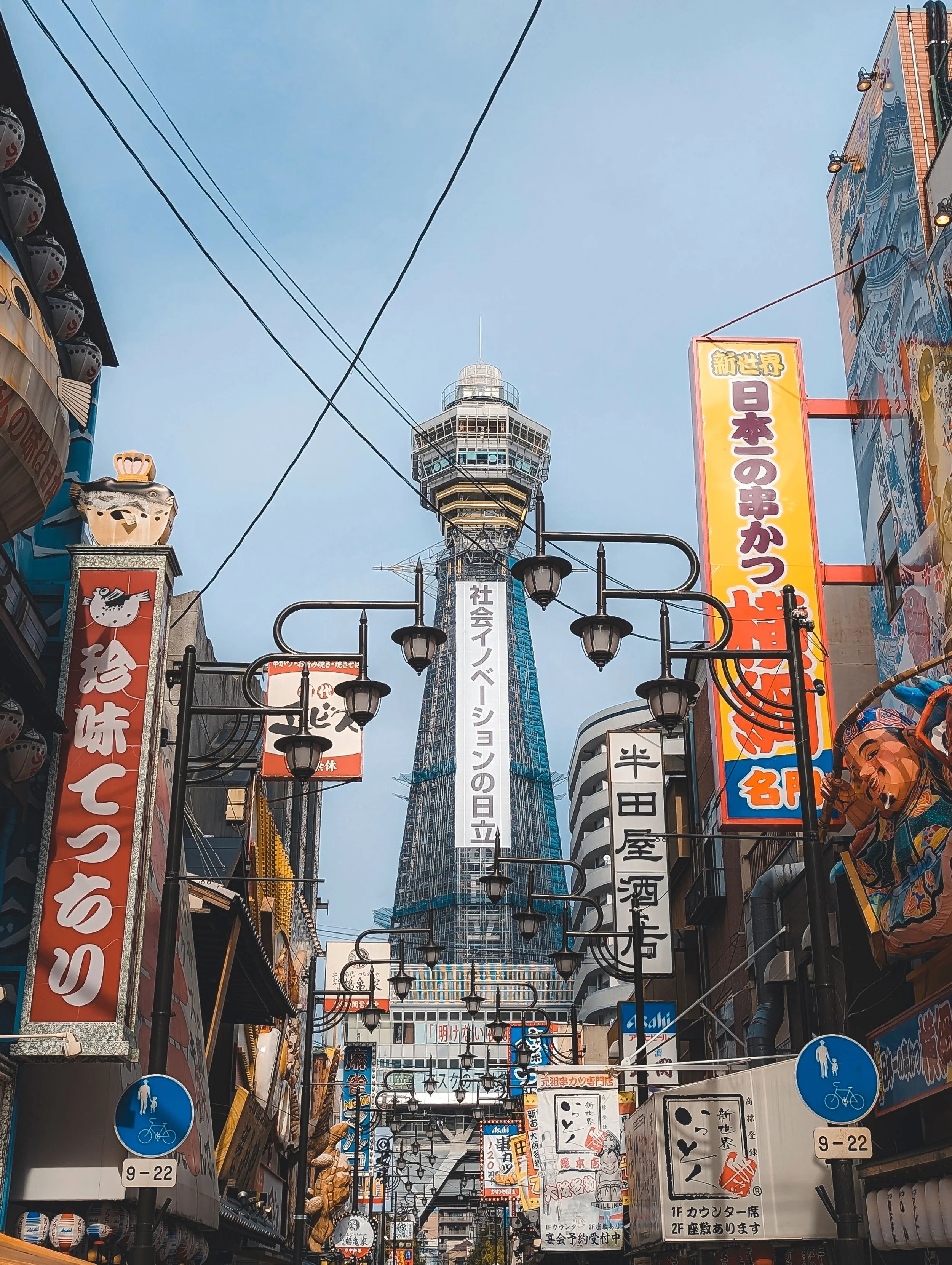 Tokyo Skytree tower visible in the background above a busy shopping street filled with colorful signs, banners, and streetlights, with pedestrians and storefronts on either side.