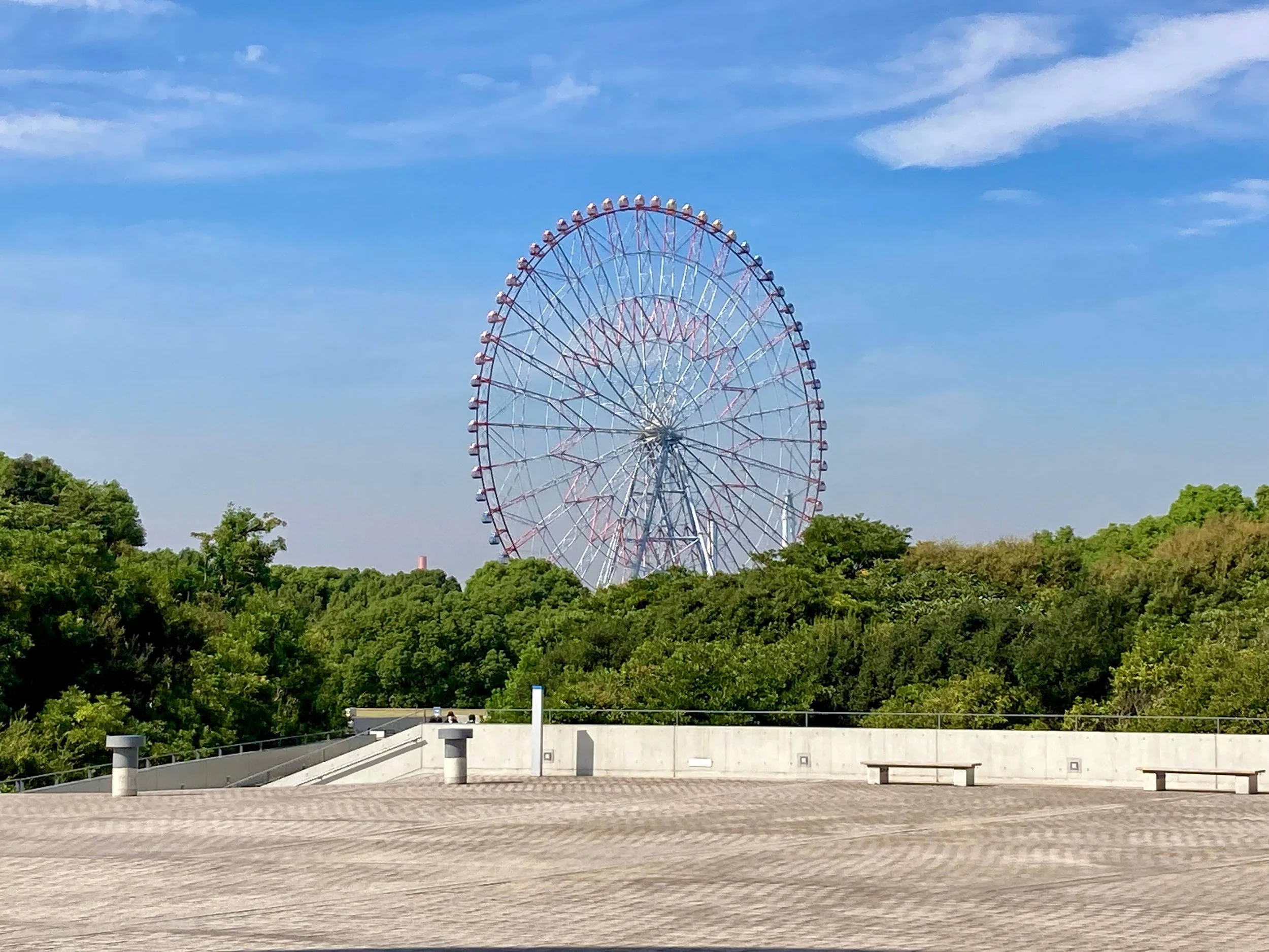 A large Ferris wheel standing against a bright blue sky with scattered clouds, surrounded by green trees, with a concrete terrace in the foreground.