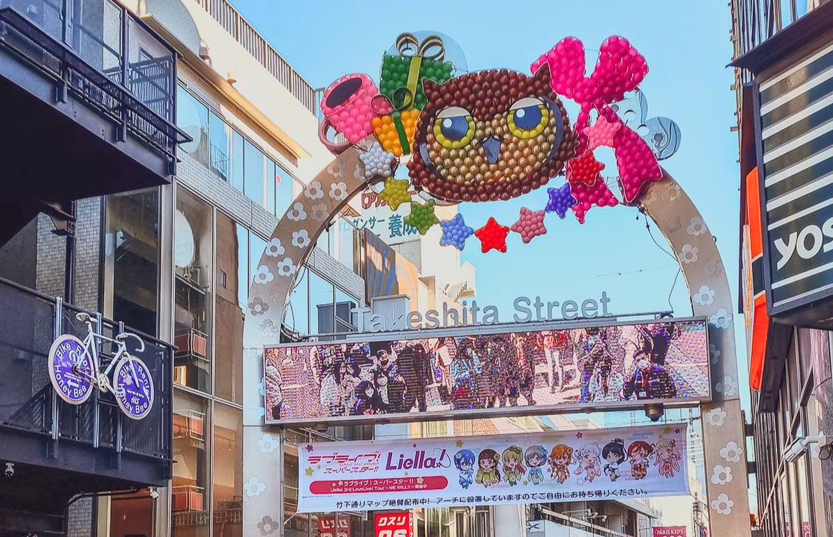 Decorative archway on Takeshita Street with colorful balloons in the shape of an owl holding gifts, a pink heart, and a star garland, with signs and buildings in the background.