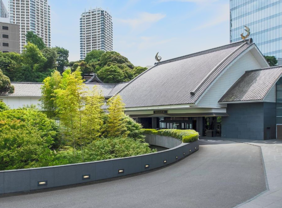 Traditional Japanese building with a curved roof and decorative finials, surrounded by lush green trees, and tall modern buildings in the background.