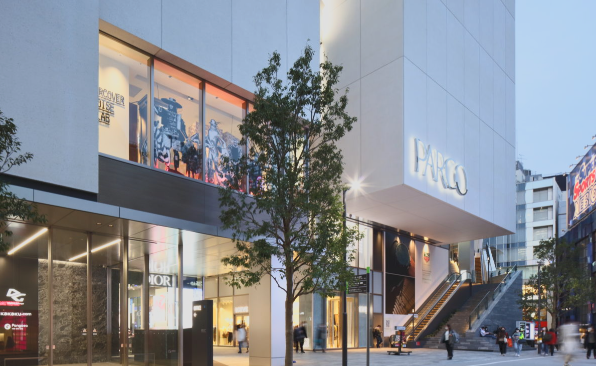 Modern shopping mall with large glass windows, illuminated signs, and people walking outside during dusk.