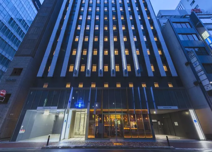 Tall modern office building with illuminated windows at night, located on a city street with neighboring buildings.