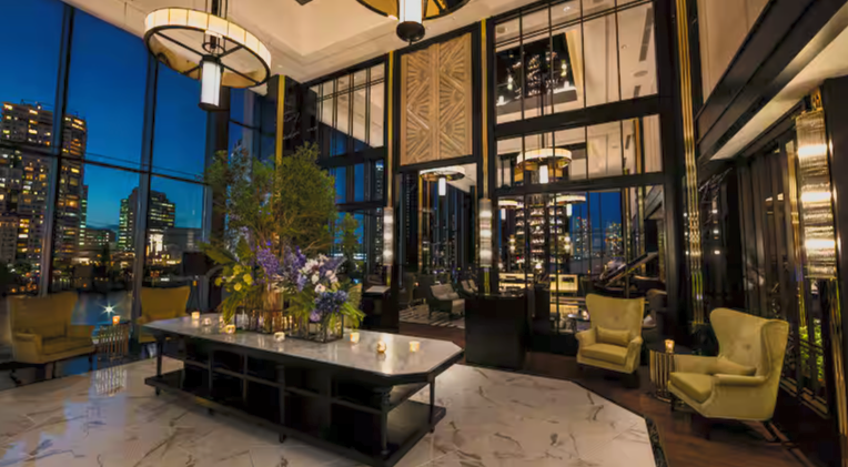 Luxury hotel lobby with large windows showing city skyline at night, modern lighting fixtures, a marble floor, and seating area with yellow armchairs and a large floral centerpiece.
