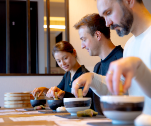 Three people preparing food at a counter, with bowls in front of them, in a kitchen or restaurant setting.