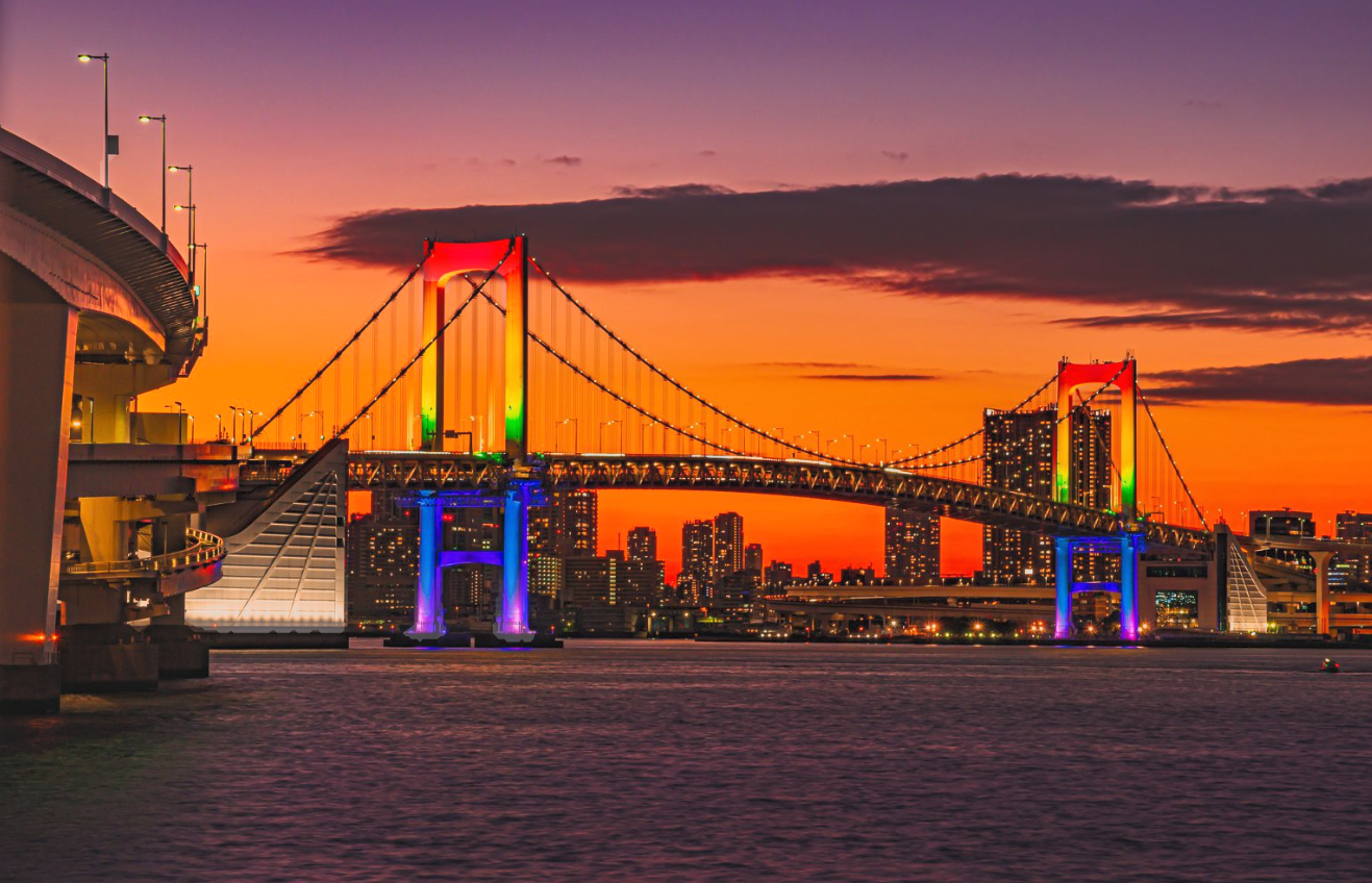 A colorful illuminated suspension bridge at sunset over a city skyline with high-rise buildings, on a body of water.