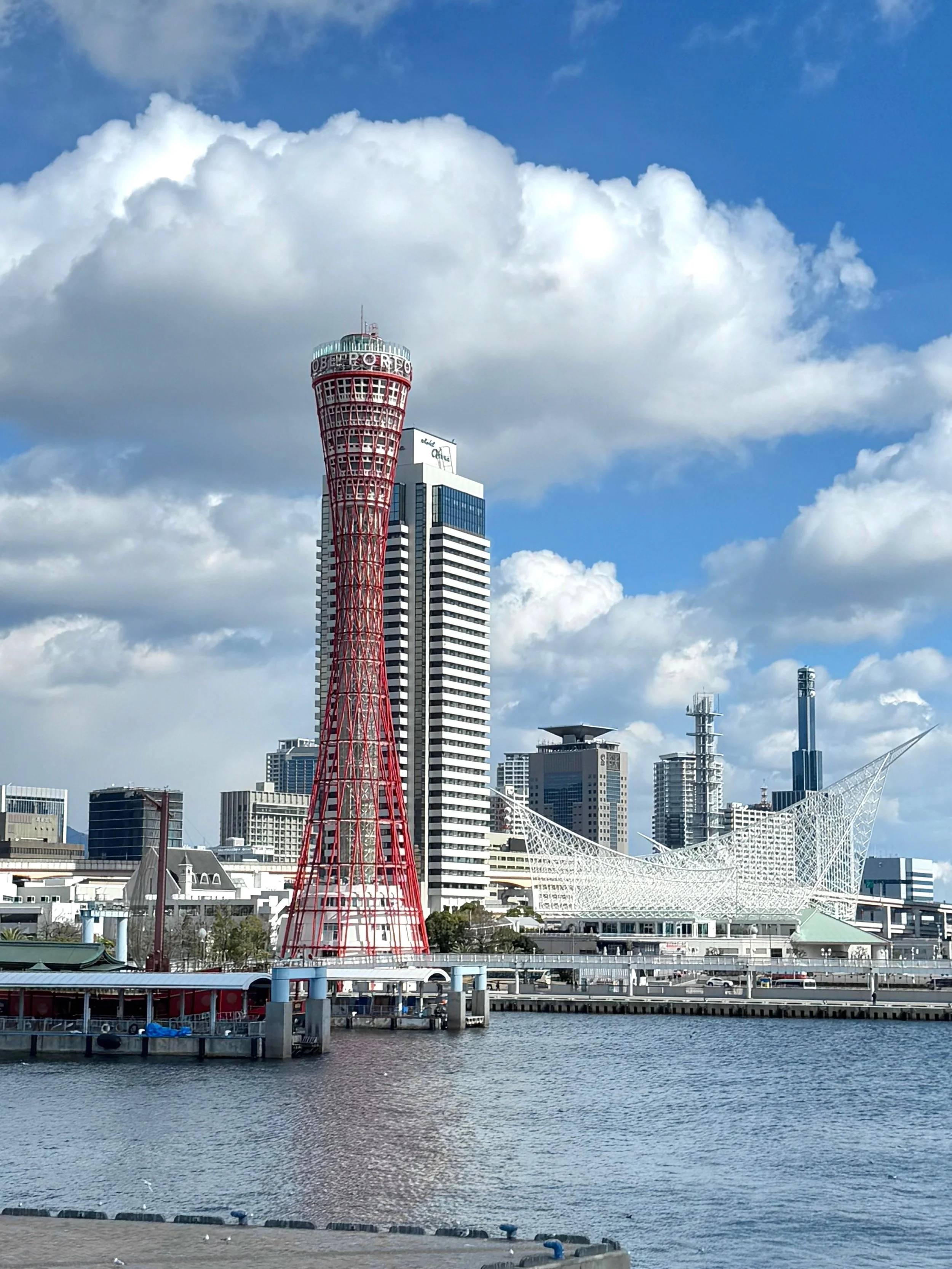 Skyline featuring the Kobe Port Tower in Japan with surrounding modern high-rise buildings, a waterfront, and a bridge.