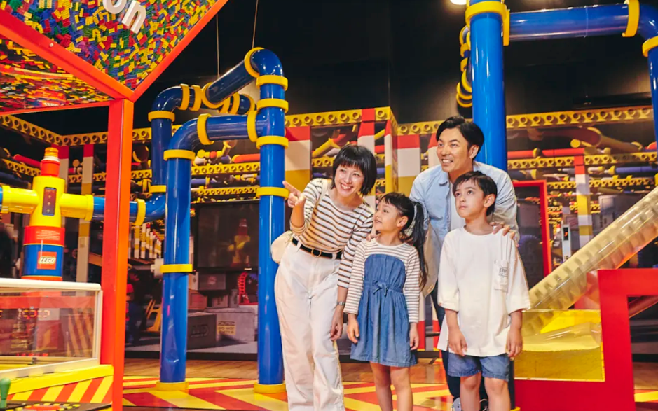 A family of four, including a woman, a man, and two children, smiling and posing inside a Lego-themed indoor play area with colorful bamboo and construction-themed structures.