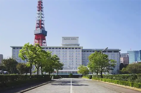 A large white hotel building with a red and white radio tower in front, surrounded by green trees and a wide paved pathway leading to the entrance.