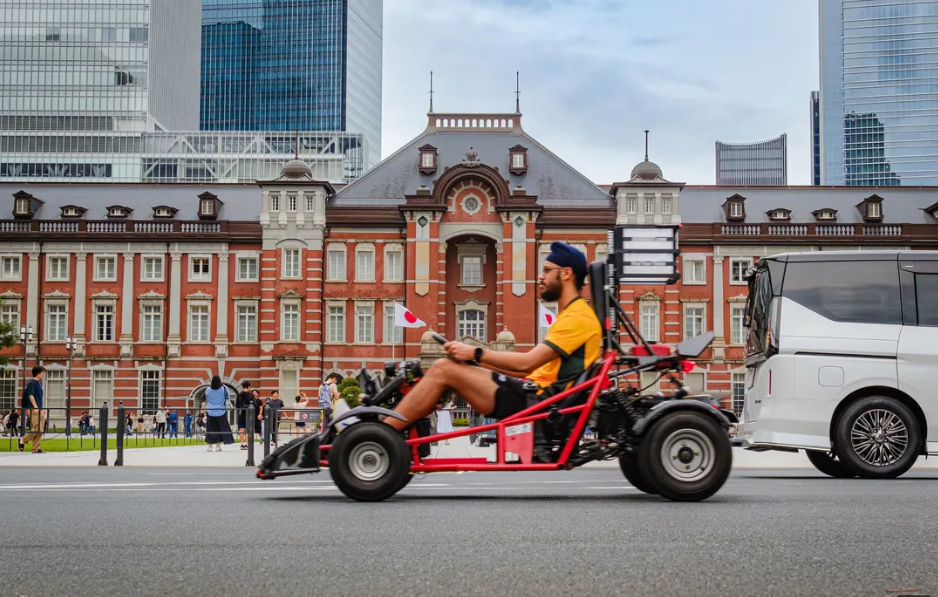 A man riding a red go-kart with a large camera on the back, traveling on a city street near a white van and historic red-brick buildings, with modern high-rise buildings in the background.