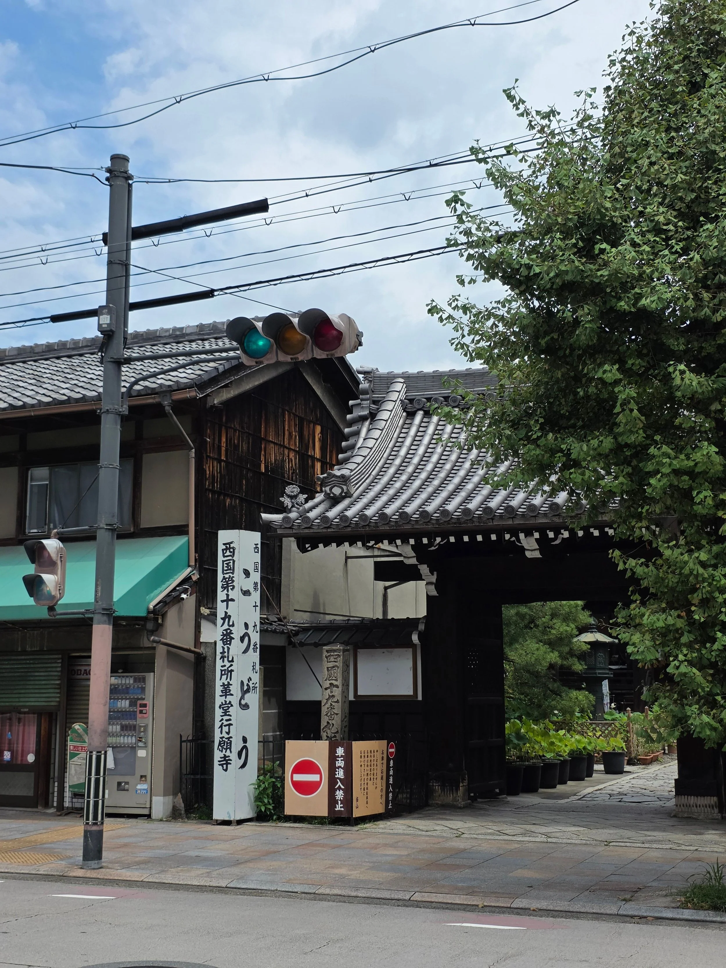 Street scene with traditional Japanese wooden buildings, a traffic light showing green, utility wires, signs with Japanese characters, and potted plants along a walkway.