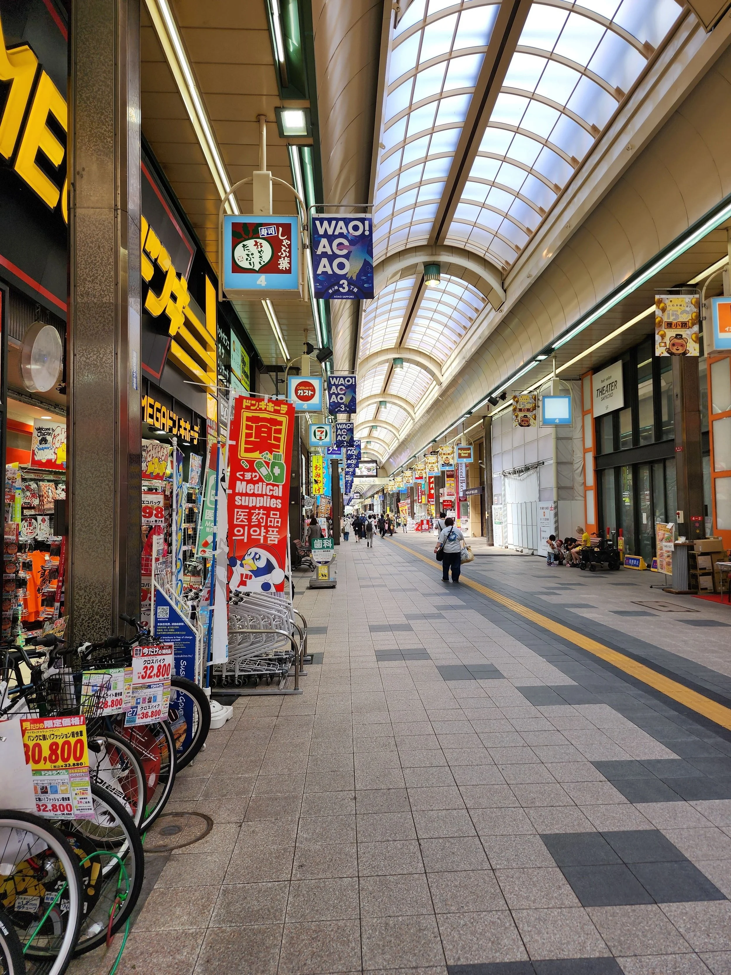 Outdoor shopping arcade with bicycles and various store signs, shoppers walking, and a covered roof with transparent panels.