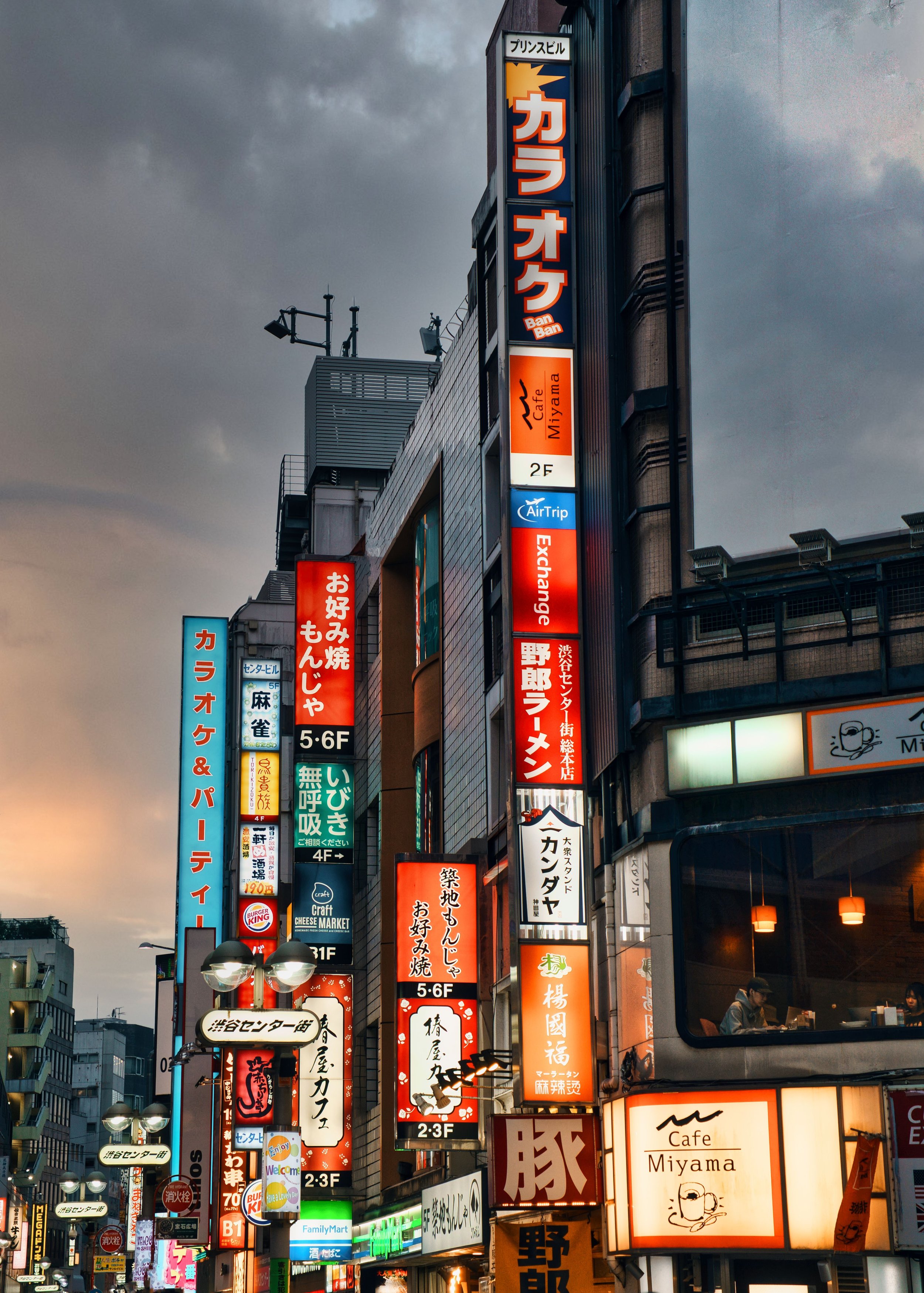 A city street at dusk in Japan, illuminated by numerous colorful neon signs in Japanese and English advertising various businesses including cafes, restaurants, and shops, with cloudy sky overhead and a mix of modern buildings.