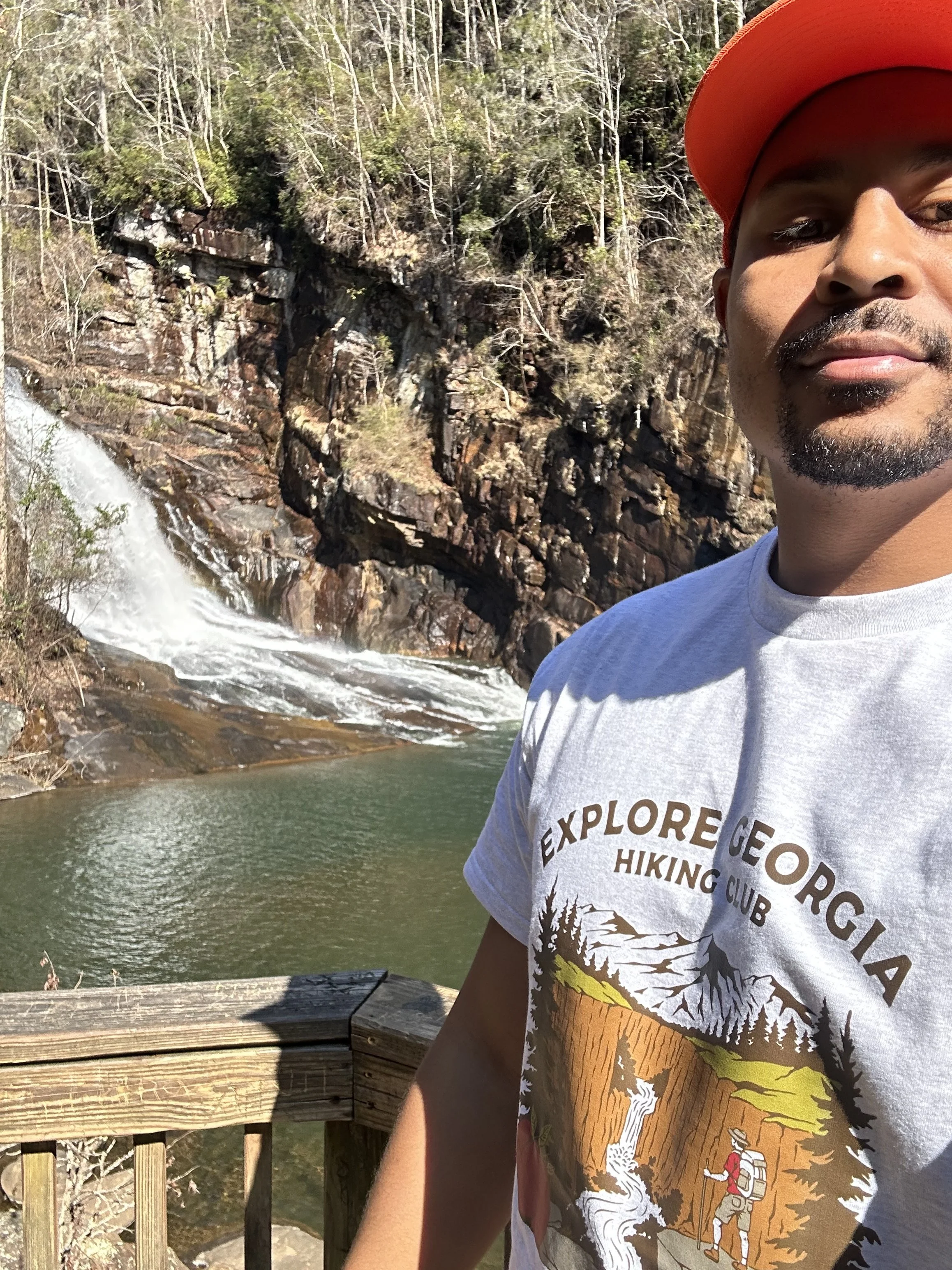 Half of a person's face and upper body wearing a gray T-shirt with mountain and outdoor scene, with a waterfall and rocky river background, standing near a wooden railing on a bright, sunny day.