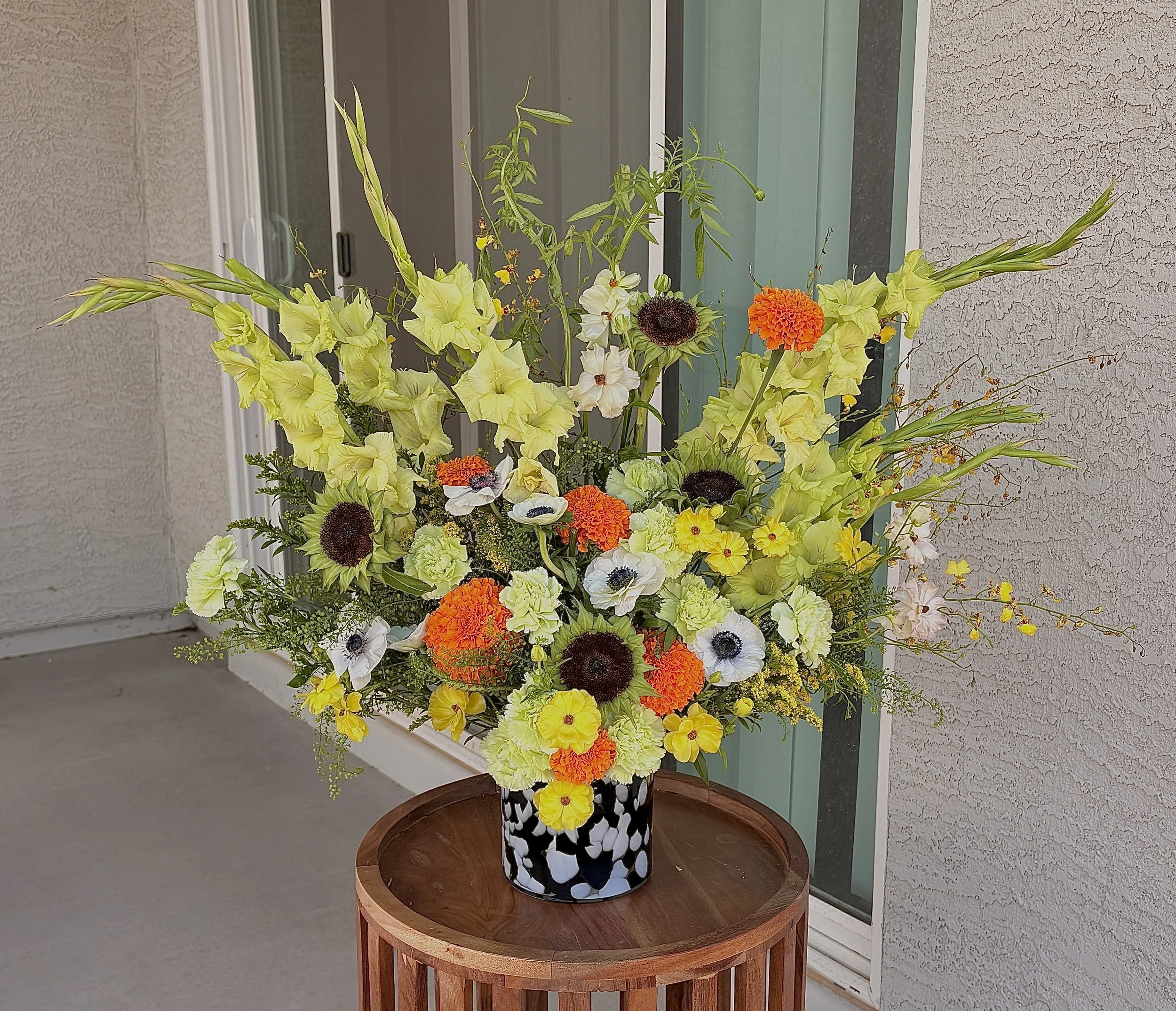 A large flower arrangement with yellow, white, orange, and dark burgundy flowers in a black and white patterned vase on a round wooden table outside near a house wall.