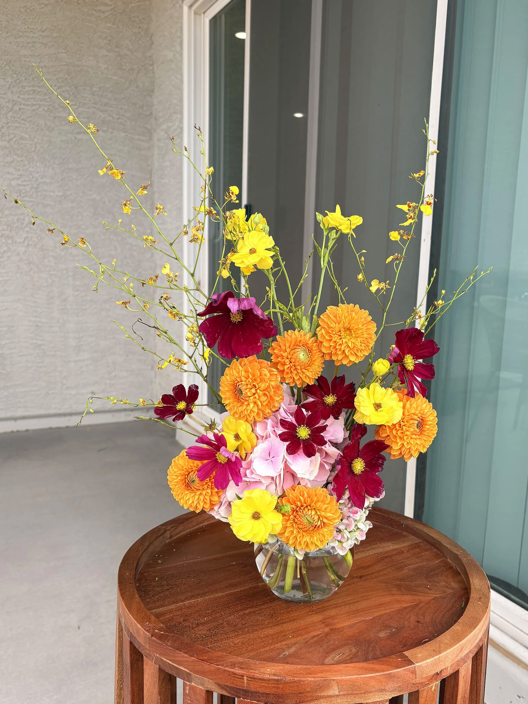 A clear glass vase filled with a colorful bouquet of various flowers, including yellow, red, orange, and pink blooms, is placed on a round wooden table near a sliding glass door.
