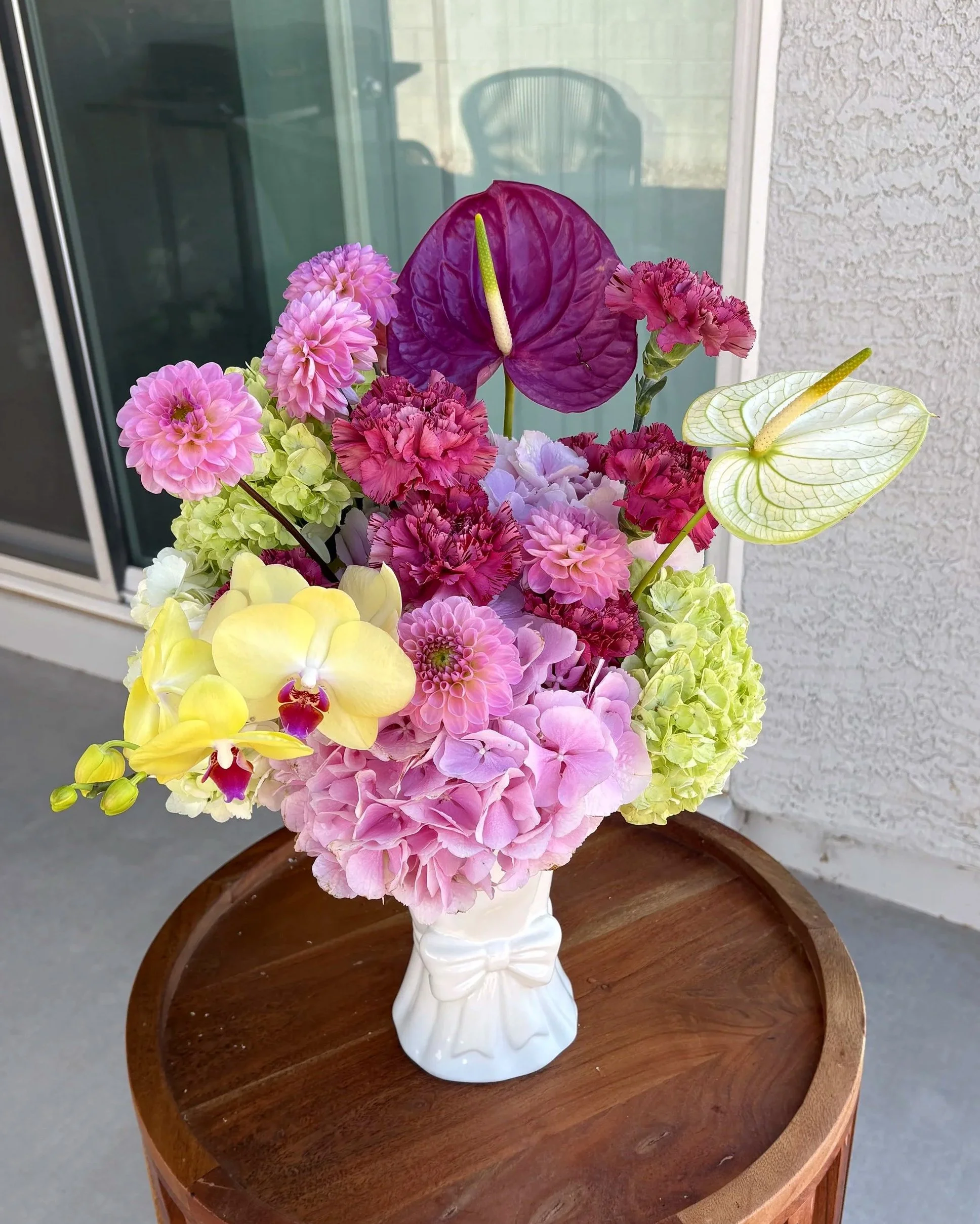 A floral arrangement in a white ceramic vase with a bow, featuring pink, purple, and yellow flowers on a wooden table outside near a sliding glass door.
