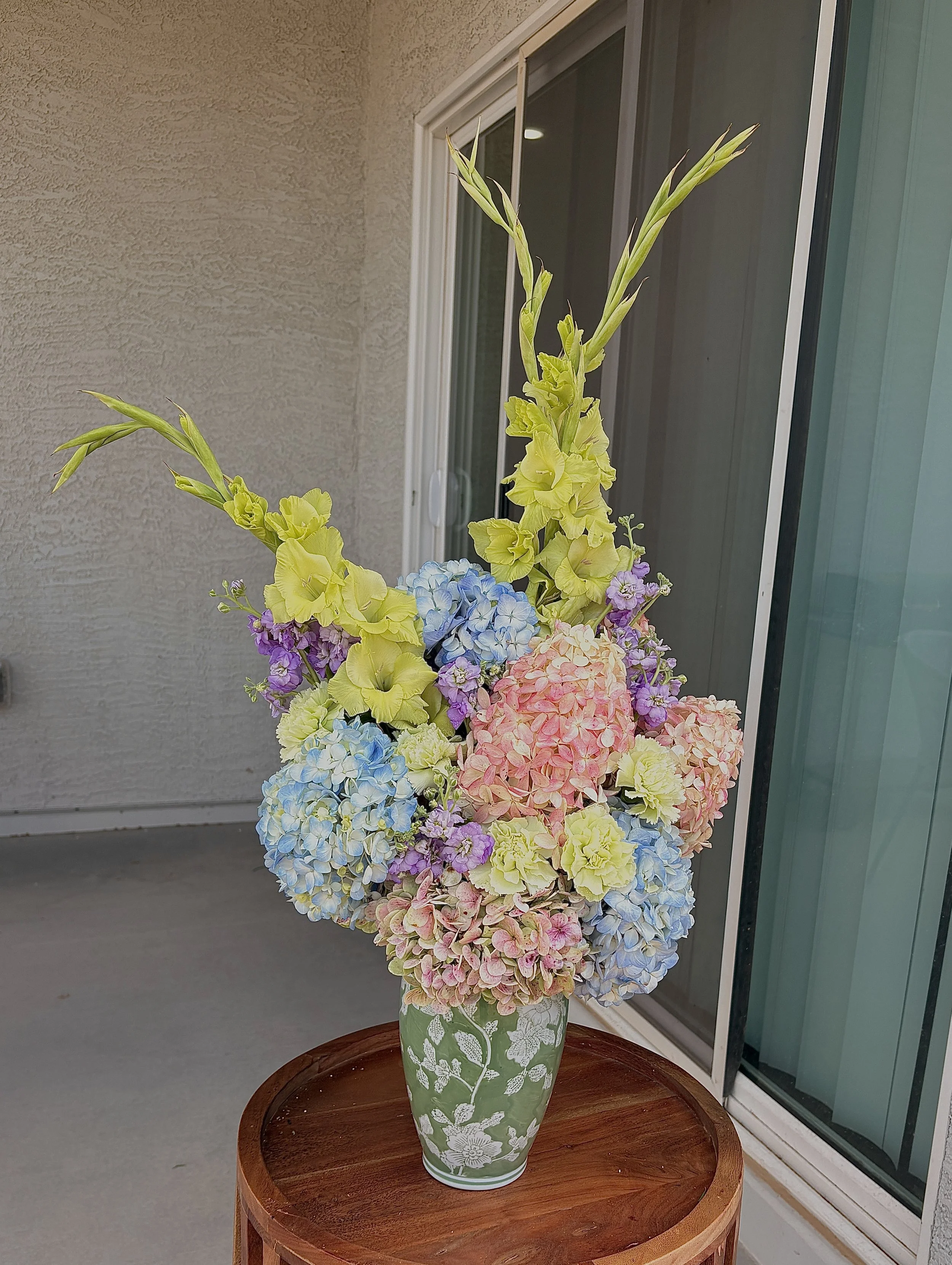 A tall green vase with white floral patterns, filled with an arrangement of colorful hydrangea and gladiolus flowers, placed on a round wooden table next to a sliding glass door.
