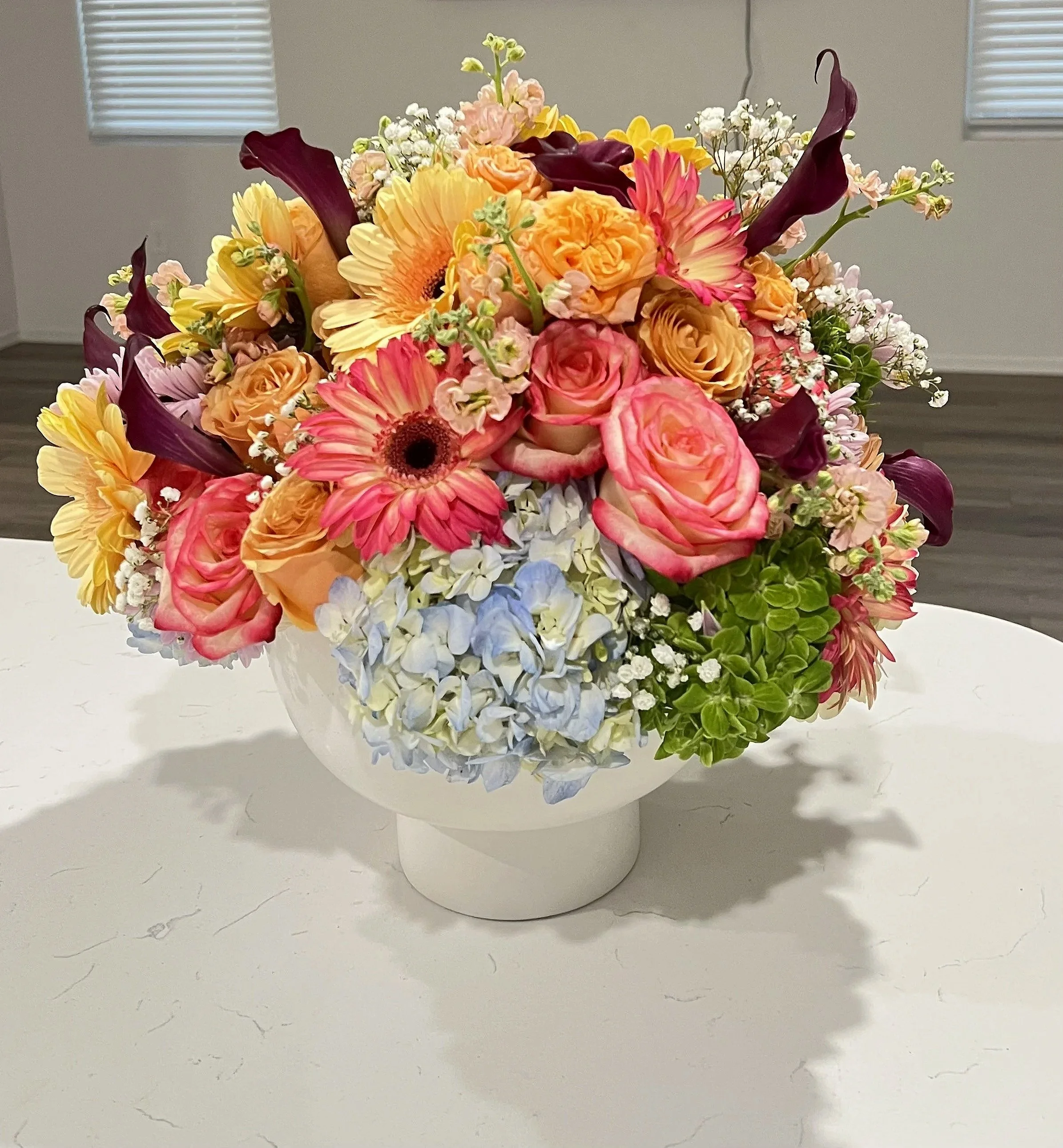 A floral arrangement in a white vase containing yellow, peach, pink, purple, and white flowers, placed on a white surface with a gray wall and window blinds in the background.