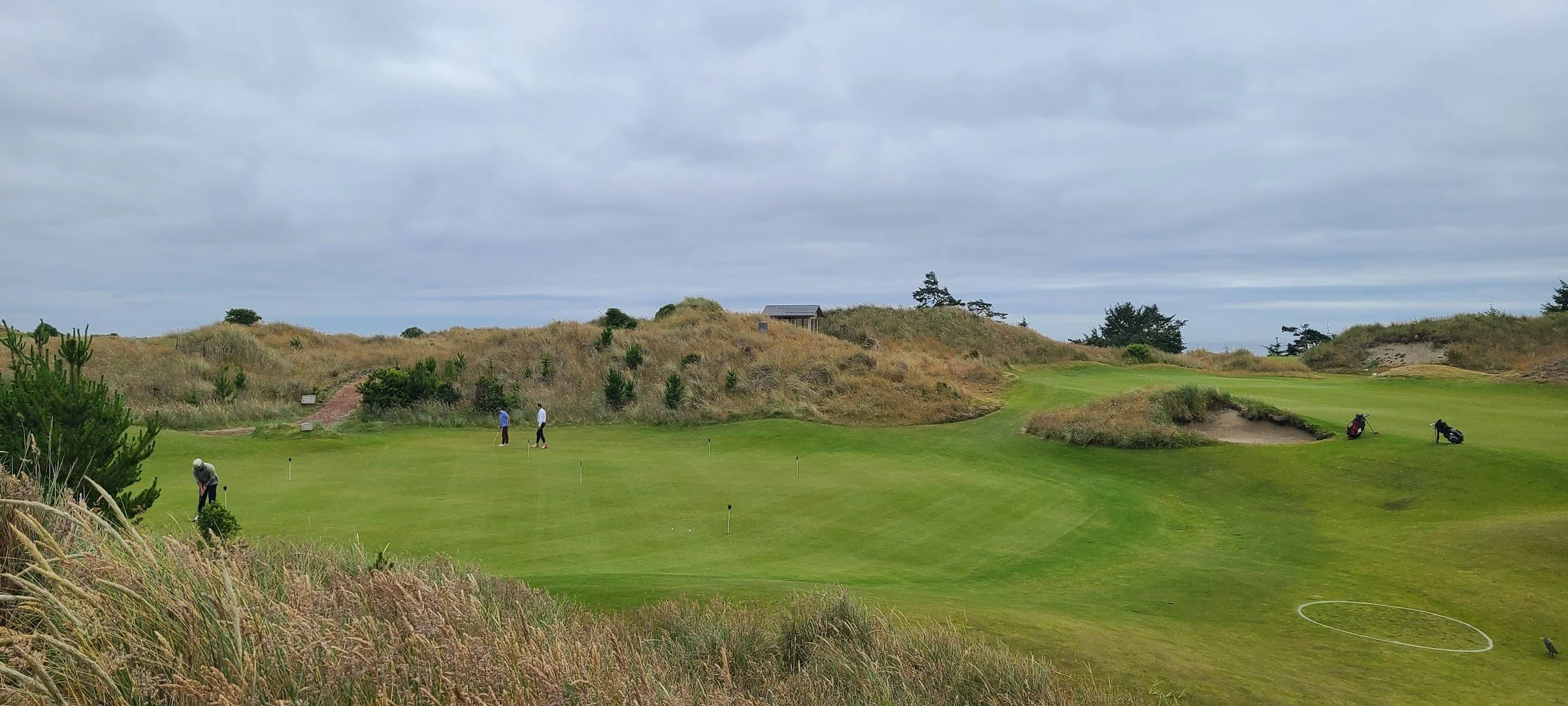 A scenic view of a golf course with players practicing their swings on the green, surrounded by sand traps and grassy hills, under a cloudy sky.
