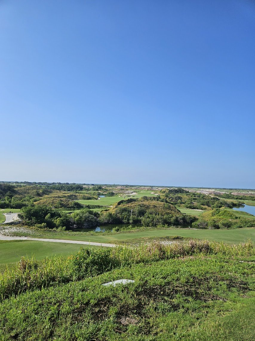 A view of a golf course with lush green grass, ponds, and trees under a clear blue sky.