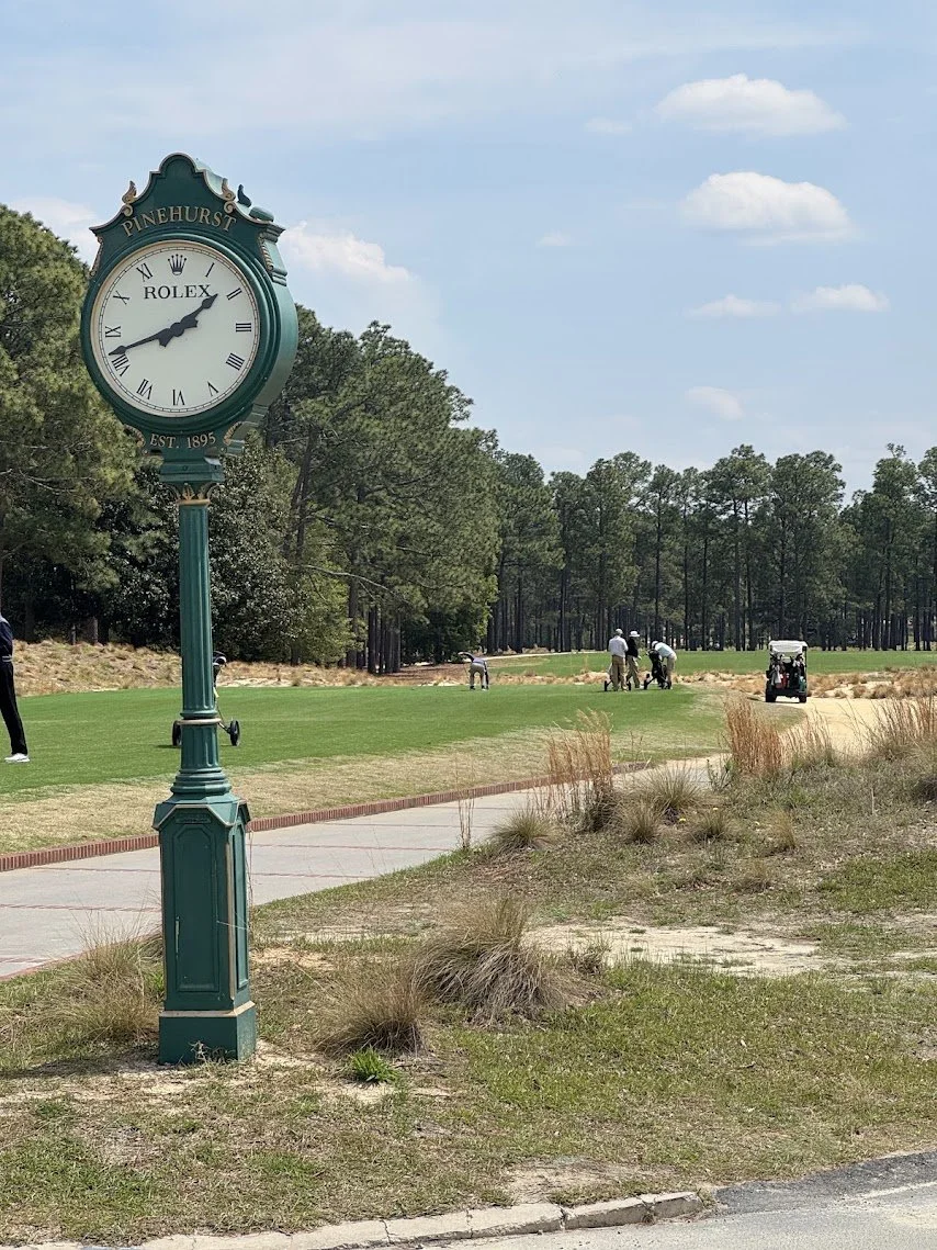 A clock with the words Pinehurst and Rolex, showing the time as 1:07, located on a golf course with people playing golf and a golf cart in the background, surrounded by trees and grassy areas.