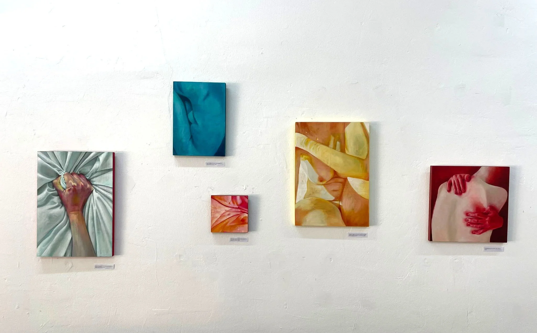 Gallery wall with six colorful paintings from Erika Clugston's series "When the Body Speaks", some featuring close-up images of hands and body parts, displayed on a white wall.